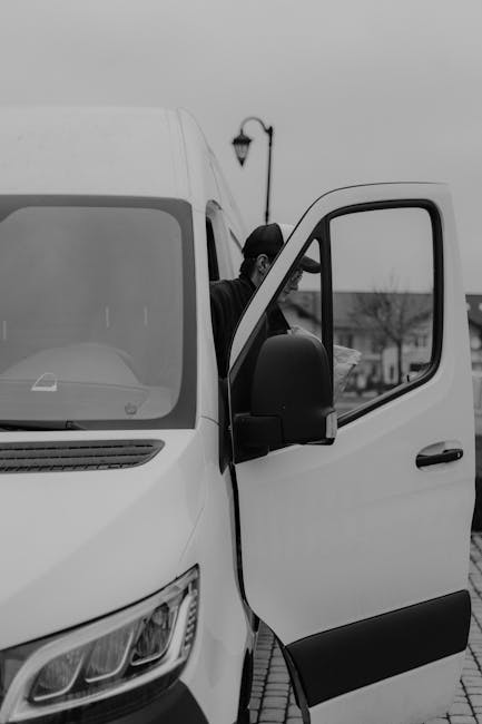 A man wearing a dark cap, grey vest, and blue long-sleeve shirt is inside the open back of a white van, which is parked outdoors near a property. He is leaning over a large cardboard box placed on a s
